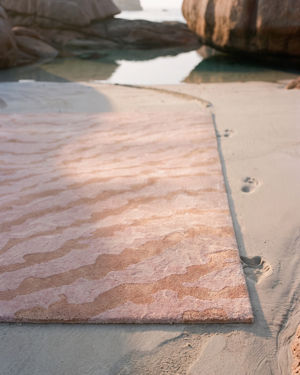 Gros plan du tapis Agata moiré rose posé sur le sable à marée basse : poil tufté main, reflets du Tencel sur le vieux rose, laine mate Rugier