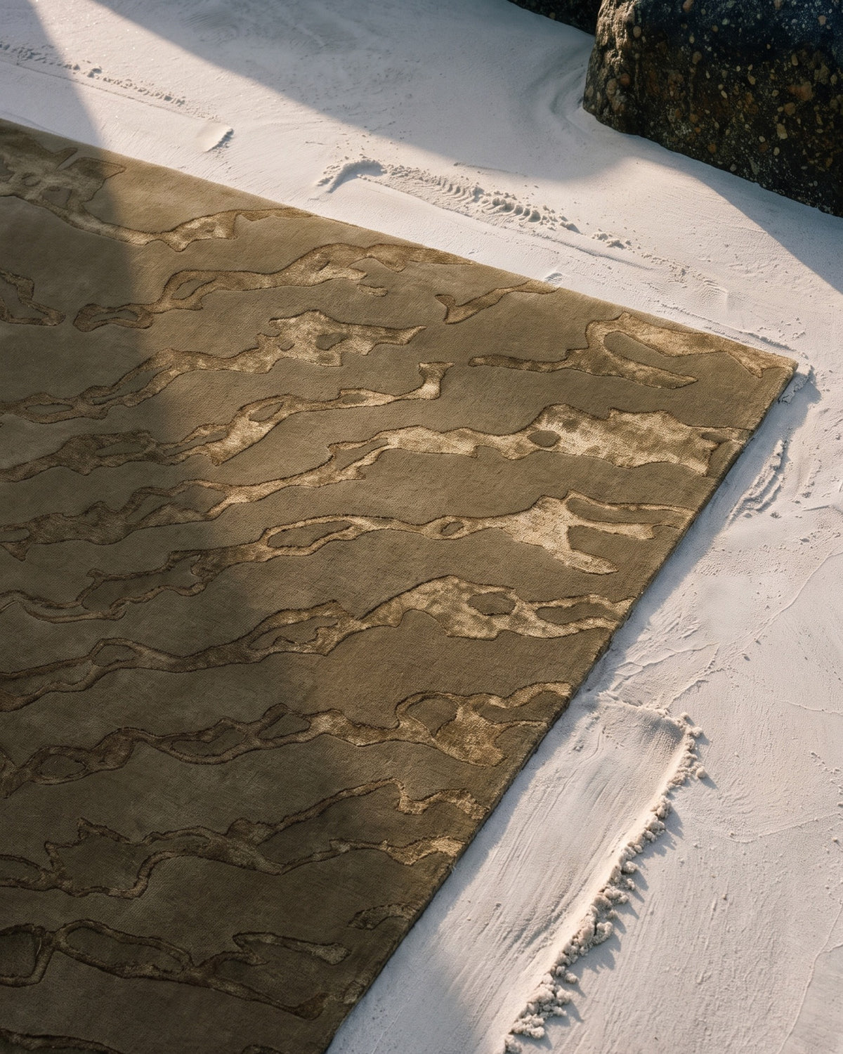 Gros plan vu du dessus du tapis Gerald moiré vert olive sur le sable, à côté d'un rocher de granit : reflets du Tencel, poil tufté main laine Rugier