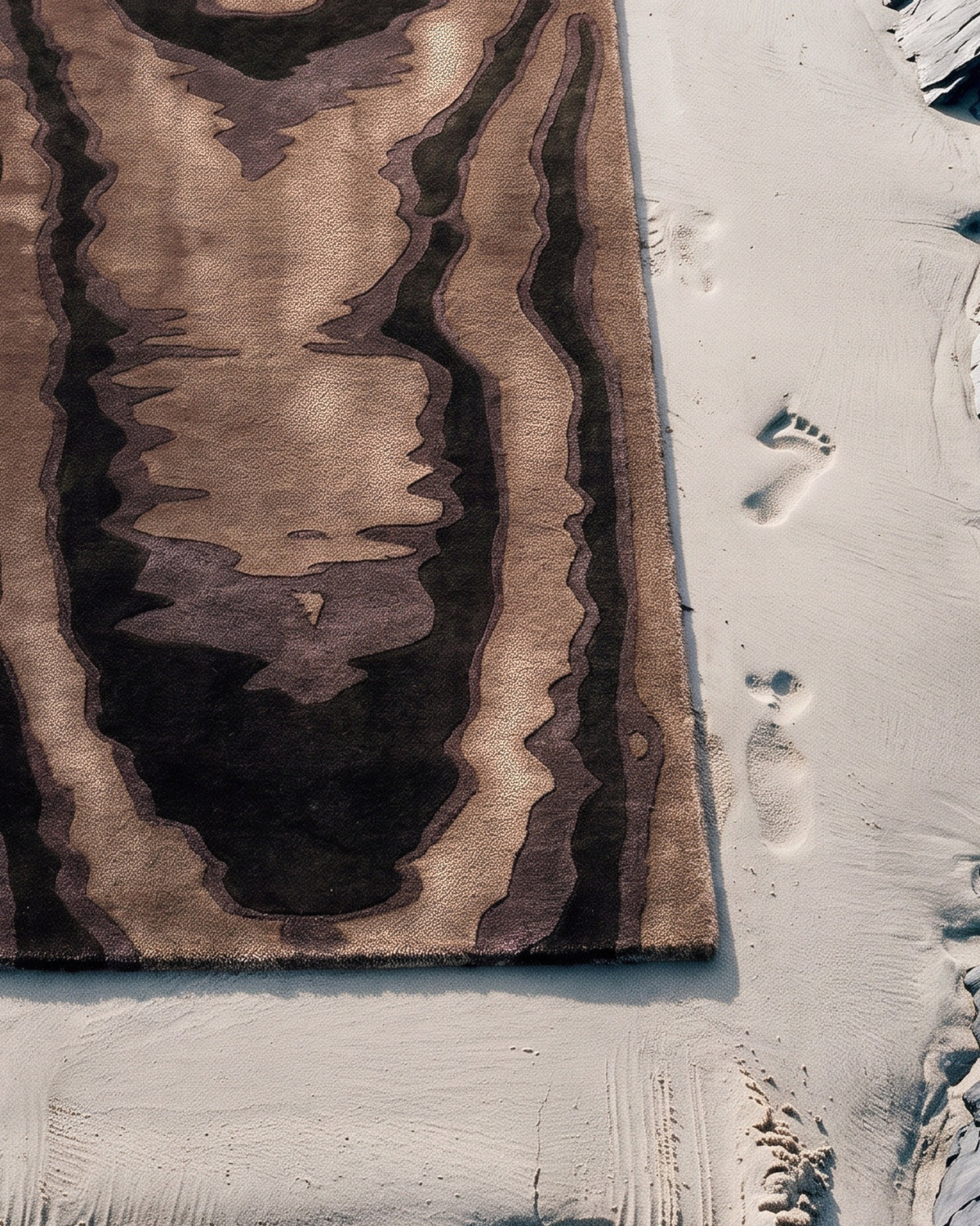 Gros plan vu du dessus du coin du tapis Harlan moiré brun sur le sable : fond tabac en Tencel satiné, veines en laine mate, tufté main Rugier